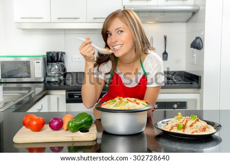 Hispanic female in modern kitchen holding spoon with food to mouth and smiling towards camera.
