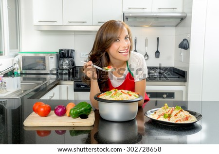 Hispanic beautiful woman cooking in modern kitchen bending over counter holding up spoon of food and smiling to camera with toungue out.