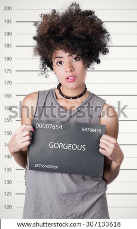 Hispanic brunette rebel model afro like hair wearing grey sleeveless shirt holding up police department board with number as posing for mugshot, careless facial expression.
