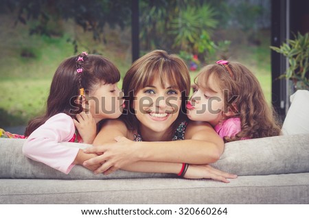 Hispanic mother in sofa with two daughters kissing her cheeks from each side, blurry garden background.