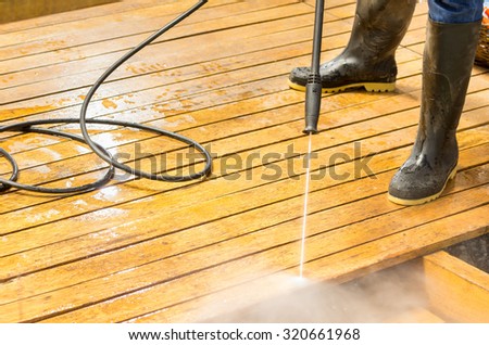 Man wearing rubber boots using high water pressure cleaner on wooden terrace surface.