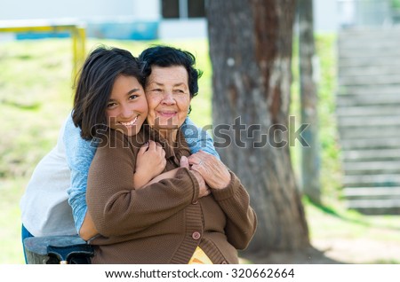 Young girl standing behind grandmother hugging and embracing in outdoors environment.