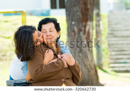 Young girl standing behind grandmother hugging and embracing in outdoors environment.