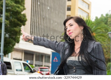Classy latina model wearing smart casual clothes walking in urban street holding arm out signalling for taxi.