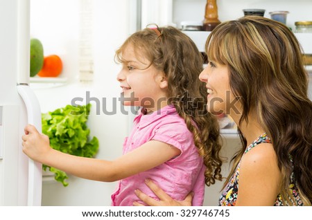 Mother holding up daughter in modern kitchen opening fridge door looking inside happily.