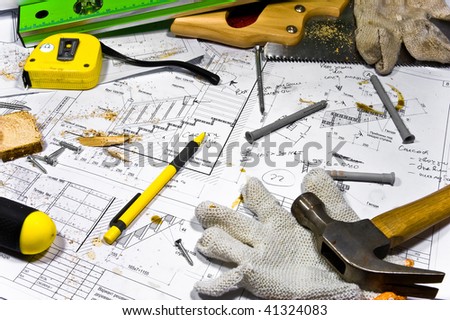 Busy hobby workbench. Different carpenter tools: saw, hummer, tape measure, level ruler, screwdriver are lying in the saw dust upon the blueprints and drawings.