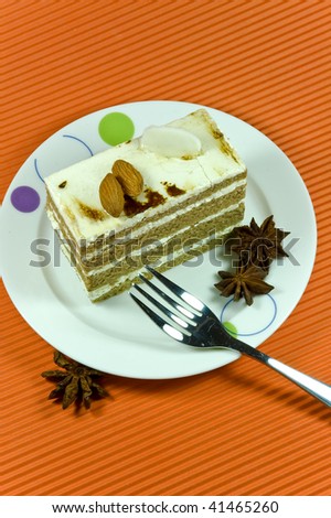Tasty and colorful brown almond cake with white cream layers. Cake is placed on the white plate along with the shiny metal fork upon the orange background.