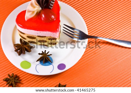 Tasty and colorful multylayer cake with chocolate decorations and red jelly. Cake is placed on the white plate along with the shiny metal fork upon the orange background.