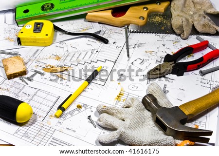 Busy hobby workbench. Different carpenter tools: saw, hummer, tape measure, level ruler, pliers and a screwdriver are lying upon the blueprints and drawings along with screws, pencil and gloves.