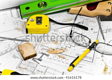 Busy hobby workbench. Different carpenter tools: saw, hummer, tape measure, level ruler, screwdriver are lying in the saw dust upon the blueprints and drawings along with screws, pencil and glasses.