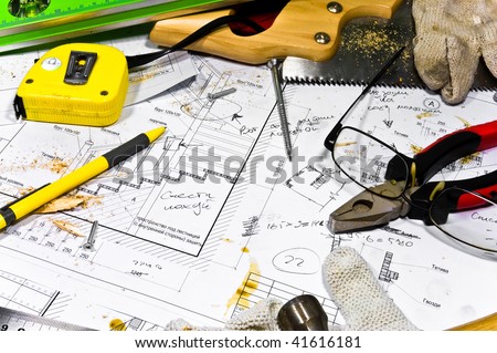 Busy hobby workbench. Different carpenter tools: saw, hummer, tape measure, level ruller and pliers are lying upon the blueprints and drawings along with screws, pencil, gloves and glasses.