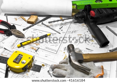 Busy hobby workbench. Different carpenter tools: drill, hummer, tape measure, level ruller and pliers are lying upon the blueprints and drawings along with screws, pencil and protective gloves.