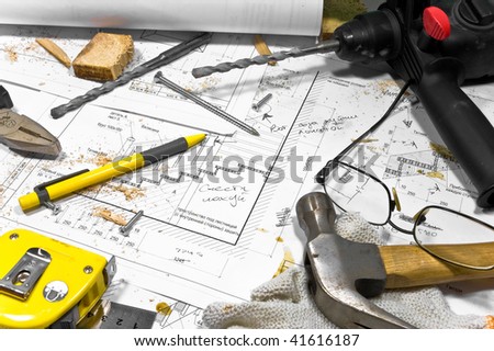 Busy hobby workbench. Different carpenter tools: drill, hummer, tape measure, level ruller and pliers are lying upon the blueprints and drawings along with screws, pencil, gloves and glasses.