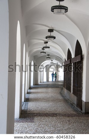 Long, white arched, arcade with light at the end and young couple holding hands, walking away at the far end