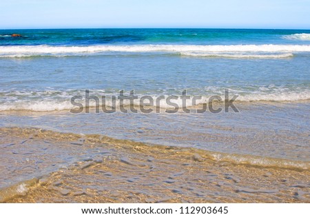Gentle waves breaking on a beach in Spain with blue sky above the horizon.  Background.
