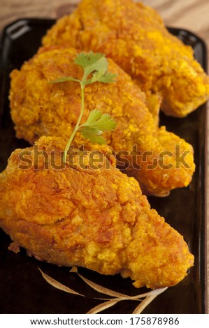 Close up of three chicken drumsticks marinated in a chili sauce then deep fried in oatmeal batter, giving a thick, crunchy covering.  Served with a garnish of coriander on a black plate