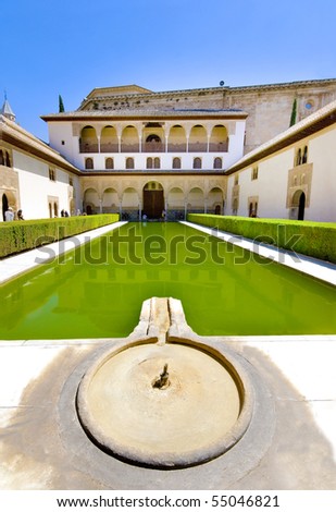 A cool Alhambra Palace fountain and pool on a hot summer's day.  The ancient red brick building has ornate mudejar arabic arches. Tourists in the background entering the palace. Granada, Spain.