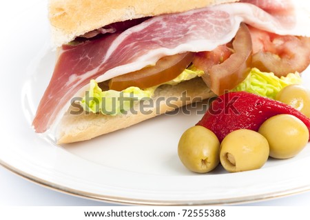 Spanish baguette sandwich full of serrano ham, tomato,  roasted piquillo pepper, olives and lettuce on a white place isolated on white.