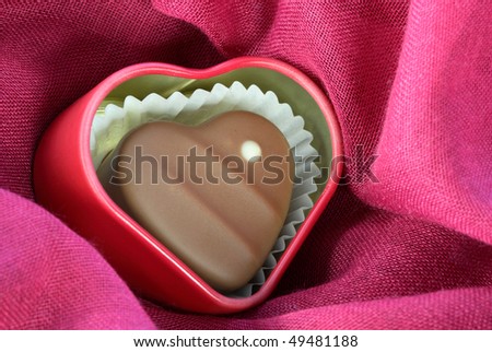 Chocolate-covered heart with a white dot on it, inside a red heart-shaped tin, on pink fabric. Horizontal shot.