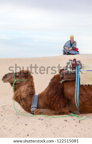 Caucasian woman sitting on sand dune in desert with camel on foreground