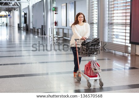 Young Caucasian woman pulling luggage hand-cart with bags along airport hall. Passenger in waiting area.