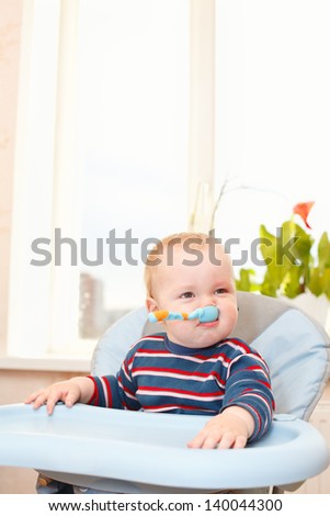 Little child sitting in chair with his spoon in mouth. Expression during eating