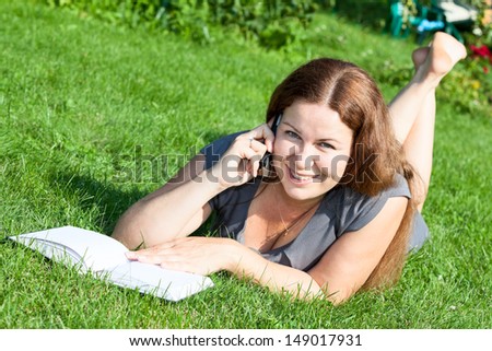 Woman on green grass reading book and speaking on phone