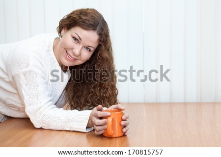 Young woman with orange mug in hands looking at camera on the table in office, copyspace