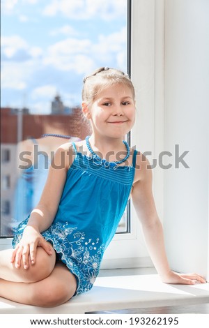 Smiling young child in blue dress sitting on window sill in sun light