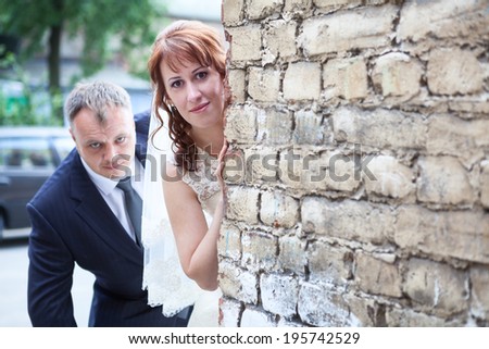 Bride and groom peek around the corner of brick wall, copyspace
