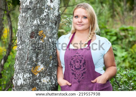 Lush Caucasian woman in apron with hands in her pockets leaning birch