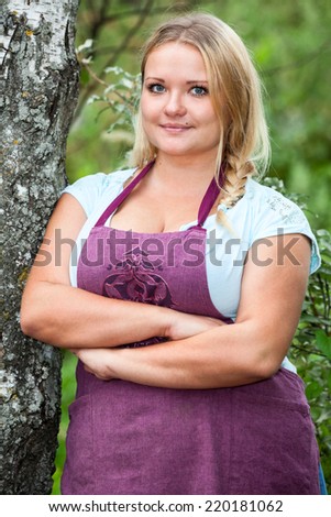 Lush Caucasian woman in apron with clasped hands leaning birch, outdoors