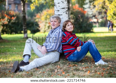 Mature couple sitting back-to-back together behind the tree in autumn park