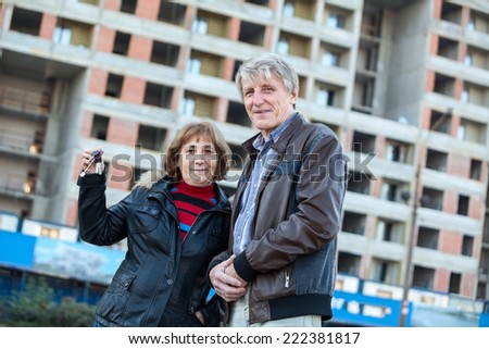 Satisfied old age couple showing house keys standing against building under construction