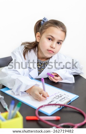 Little girl in a doctor white coat sitting at the table and looking at camera