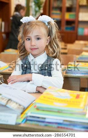 First-form cute schoolgirl sitting at school desk in classroom, looking at camera