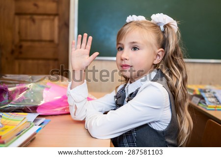 First grade schoolgirl in uniform rising hands at school desk, side view