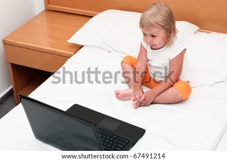 A small child is sitting with a laptop on the bed and looking at screen