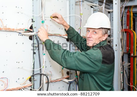 Senior Caucasian electrician working in white hard hat with cables and wires. Screwdriver in hand, looking at camera