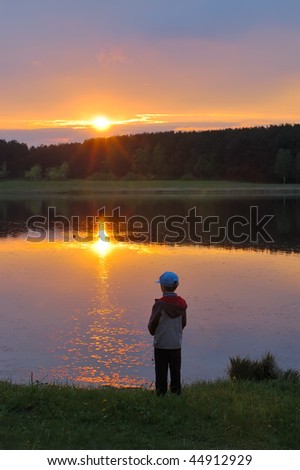 The boy stands on the shore of the lake, looking at the setting sun.