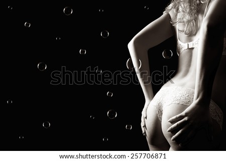 Beautiful back of a woman wearing white panties and bra in front of dark studio  background, monochrome photo with copy space and soap bubbles on the left side of the image