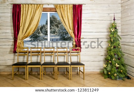 A room in a country village yellow wooden house just before Christmas and New Year. Christmas tree with toys is at the wall. Chairs for the guests at the window. Outside the window the winter forest.