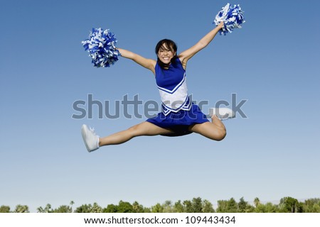 Full length of an excited young cheerleader jumping with pom-poms against sky