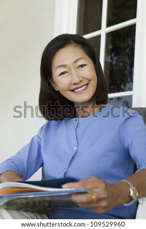 Portrait of a happy woman on chair holding magazine in front of house