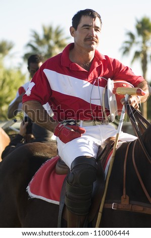 Polo player sitting on horse while holding helmet and polo stick in hand