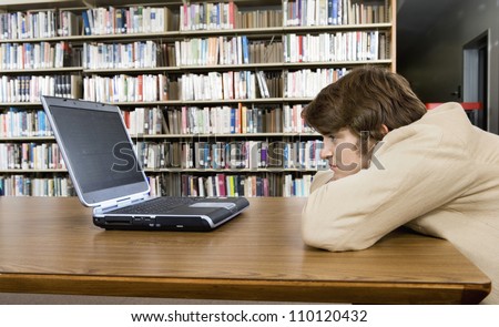 Side view of a bored male college student sitting at library desk looking at laptop