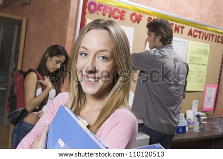 Portrait of a happy female student in college office clutching folder