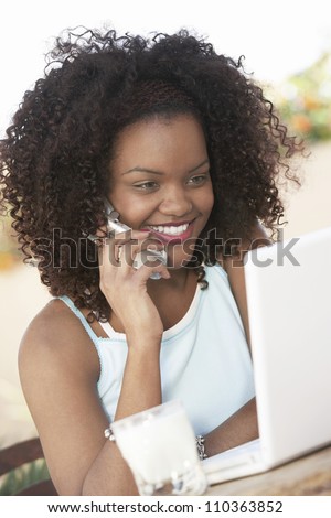 Happy young African American woman with laptop using cell phone