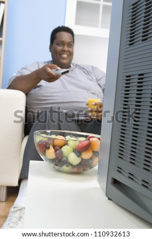 African American man drinking juice while watching television with a bowl of fruit in the front