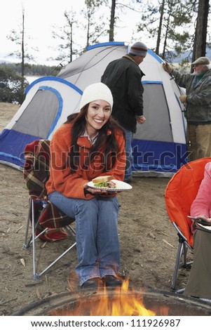Happy woman having food at campfire with man at tent in background